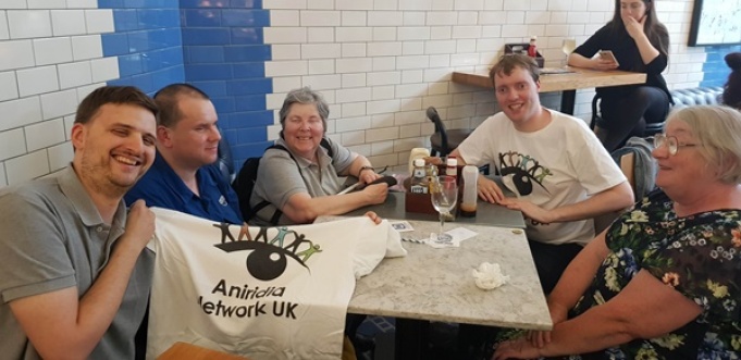 5 people around a table in London Victoria holding up a Aniridia Network UK t-shirt